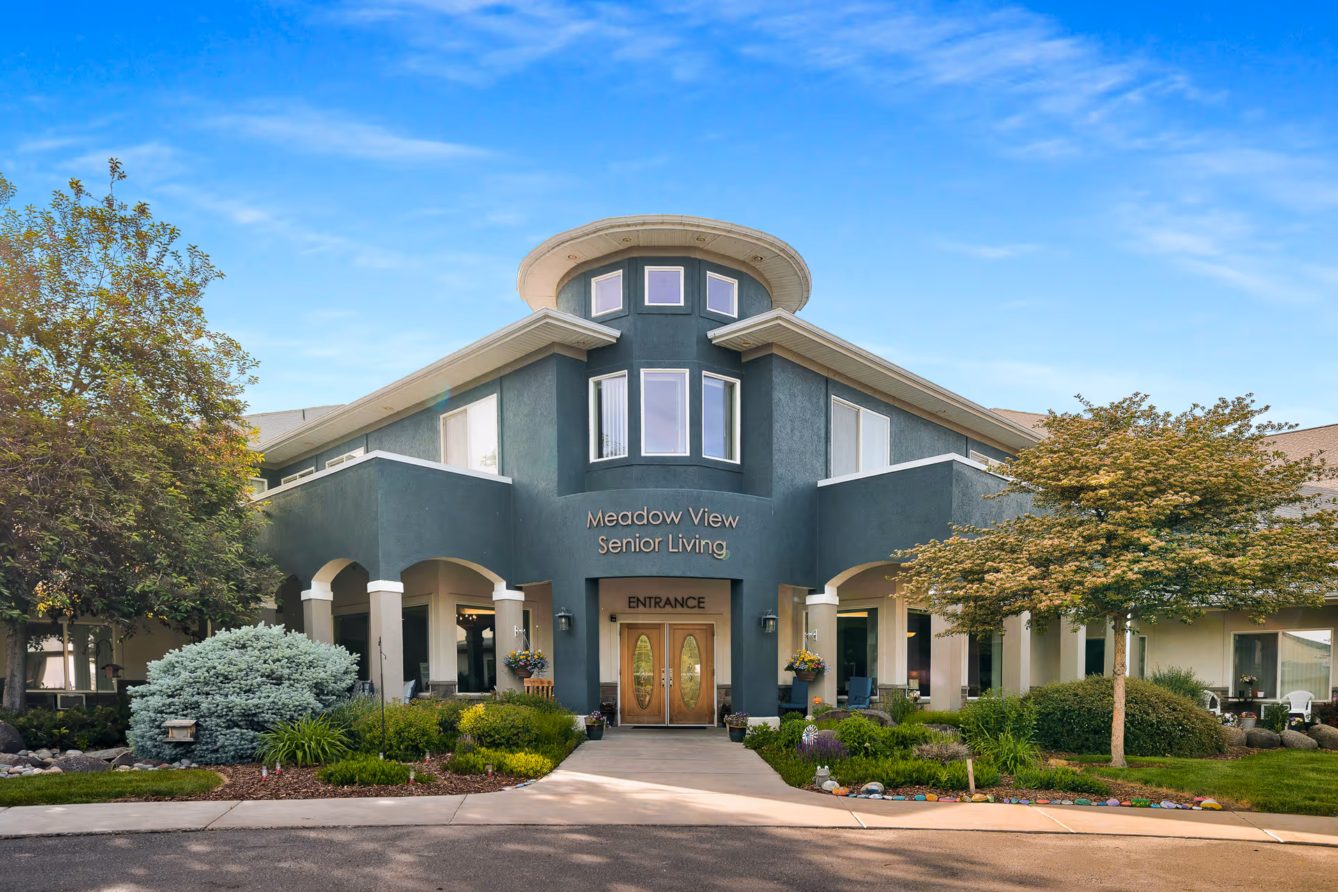 Front exterior view of Meadow View Senior Living facility with a modern two-story building, large windows, double wooden entrance doors, and landscaped greenery including trees and bushes under a clear blue sky.