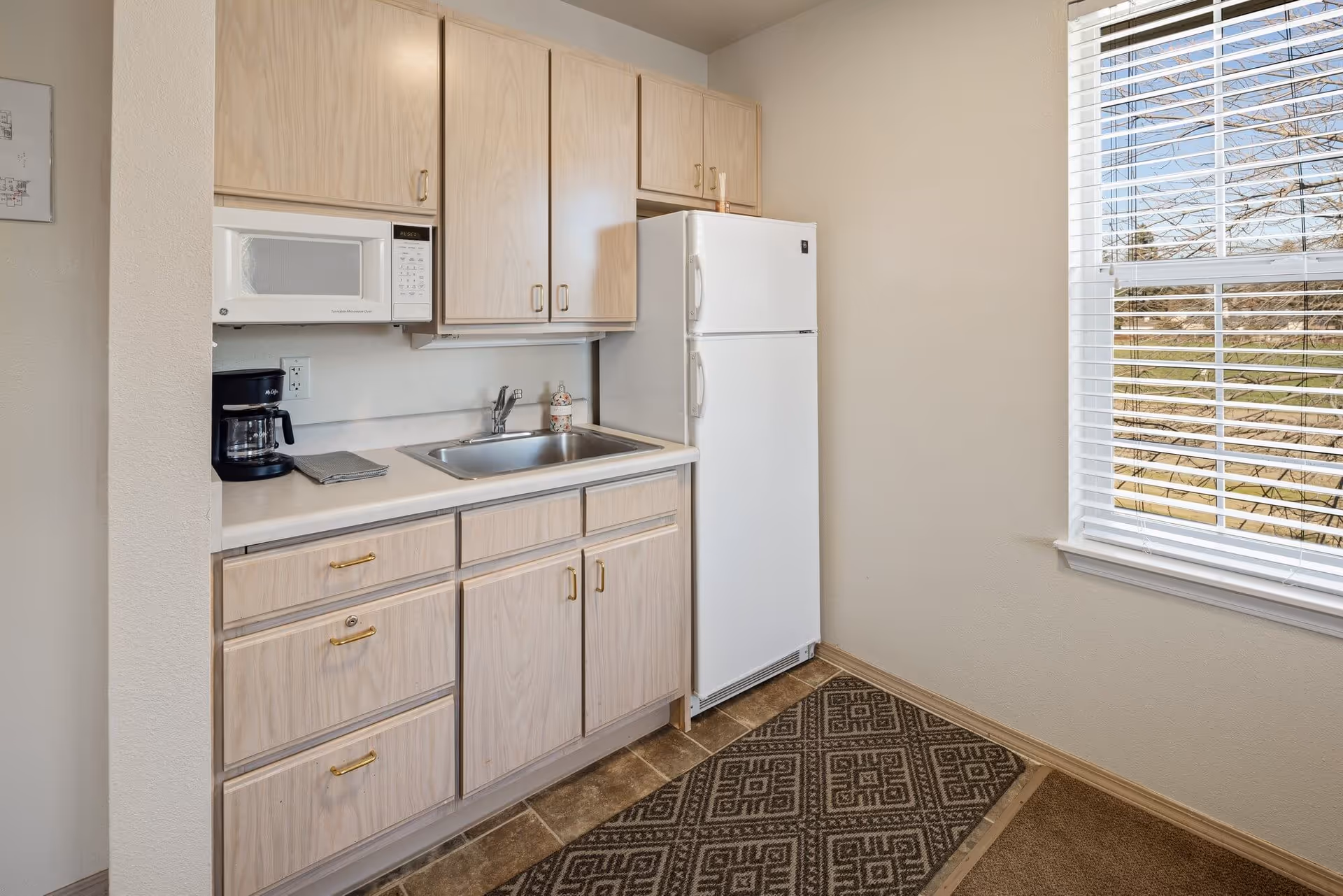 Small kitchen area with light wood cabinets, a white microwave, a coffee maker, a stainless steel sink, and a white refrigerator. There is a window with white blinds on the right side letting in natural light, and a patterned rug on the floor.
