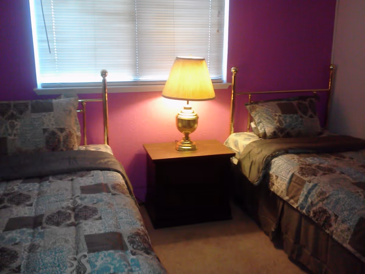 A bedroom with two twin beds separated by a wooden nightstand with a brass lamp on it. The beds have patterned bedspreads in shades of blue, brown, and beige. The wall behind the beds is painted purple, and there is a window with closed blinds above the nightstand.