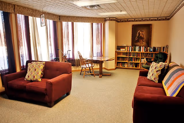 A cozy living room with two maroon sofas, each with patterned pillows. A wooden table with a chair is positioned near large windows with sheer curtains. A bookshelf filled with books and a framed picture of Jesus Christ hang on the wall. The room has beige walls and carpeted flooring.