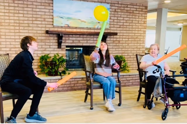Three people sitting on chairs in a room with a brick fireplace and plants. They are playing with colorful foam bats and a yellow balloon. One person is elderly and using a walker.