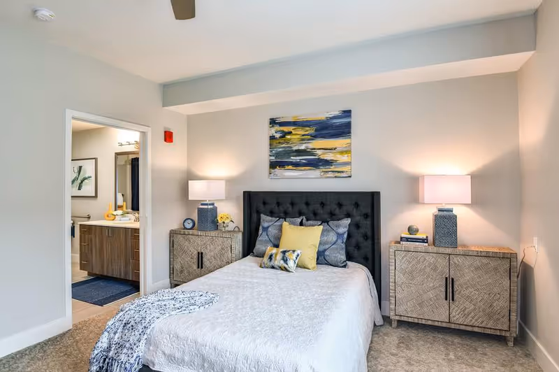 Bright bedroom with a tufted dark headboard, decorative pillows on the bed, matching nightstands with lamps, and an open door to a bathroom.