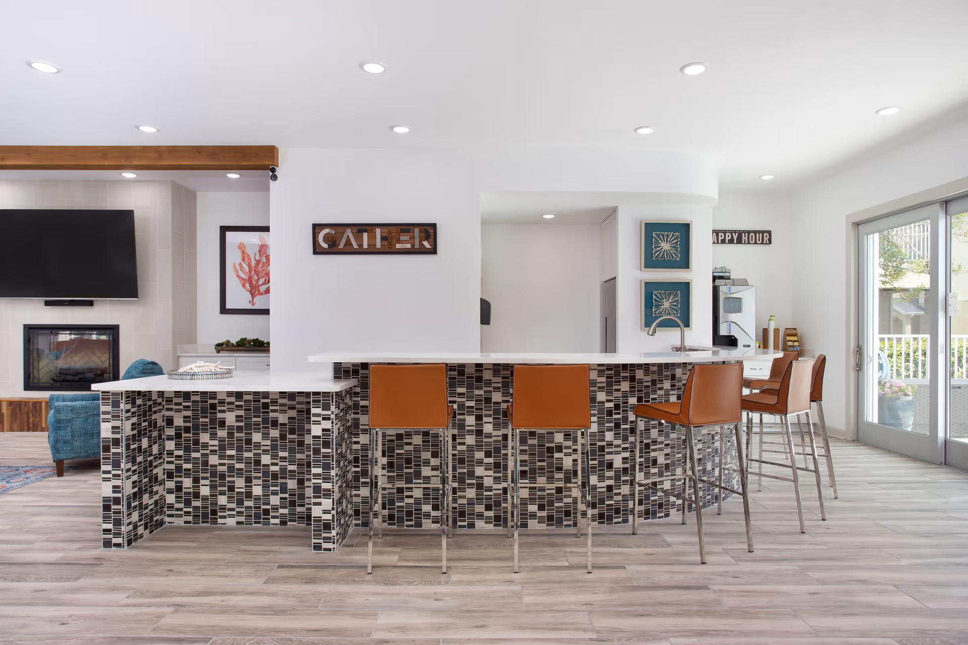 Modern senior living facility common area with a tiled bar counter and four brown bar stools. The room features light wood flooring, white walls, recessed ceiling lights, a wall-mounted TV above a fireplace, and sliding glass doors leading outside. Decorative signs reading 'GATHER' and 'HAPPY HOUR' are visible on the walls.