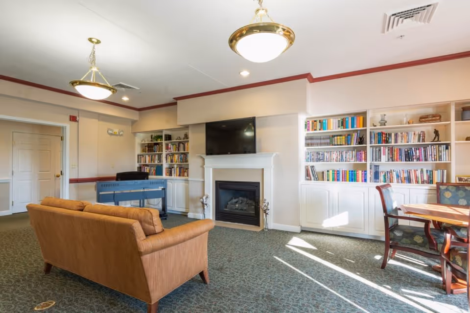 A cozy living room area in Smith Farms Manor featuring a brown sofa, a fireplace with a mounted flat-screen TV above it, built-in white bookshelves filled with books on either side of the fireplace, a wooden table with chairs near a window letting in sunlight, and a blue piano against the far wall.