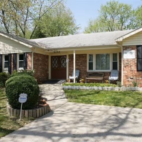 Front exterior view of a single-story brick building with a white roof and a small porch area. The entrance has double doors with glass panels, flanked by two chairs and a bench. There is a small garden with green bushes and a white picket fence in front of the porch. Trees with green leaves are visible in the background.