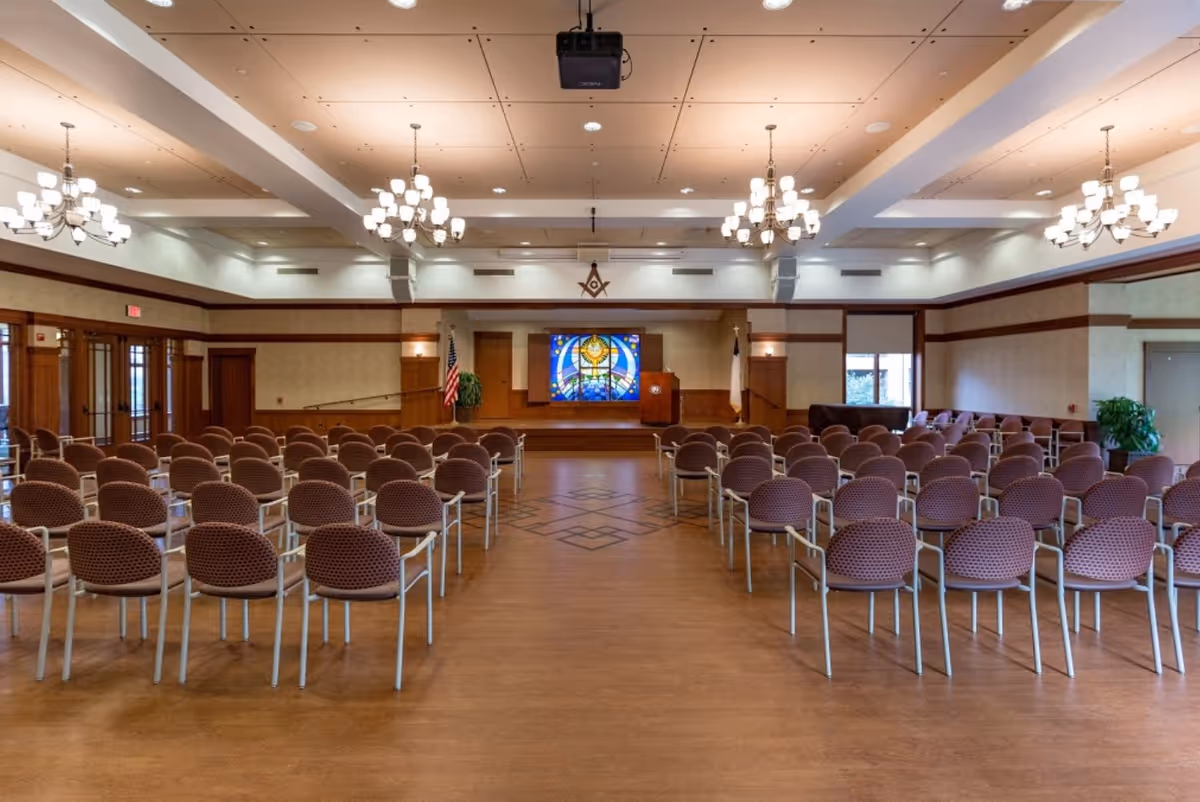 A large meeting or event room with rows of empty chairs facing a stage with a podium and a colorful stained glass window. The room has chandeliers hanging from the ceiling, wood paneling, and flags on either side of the stage.