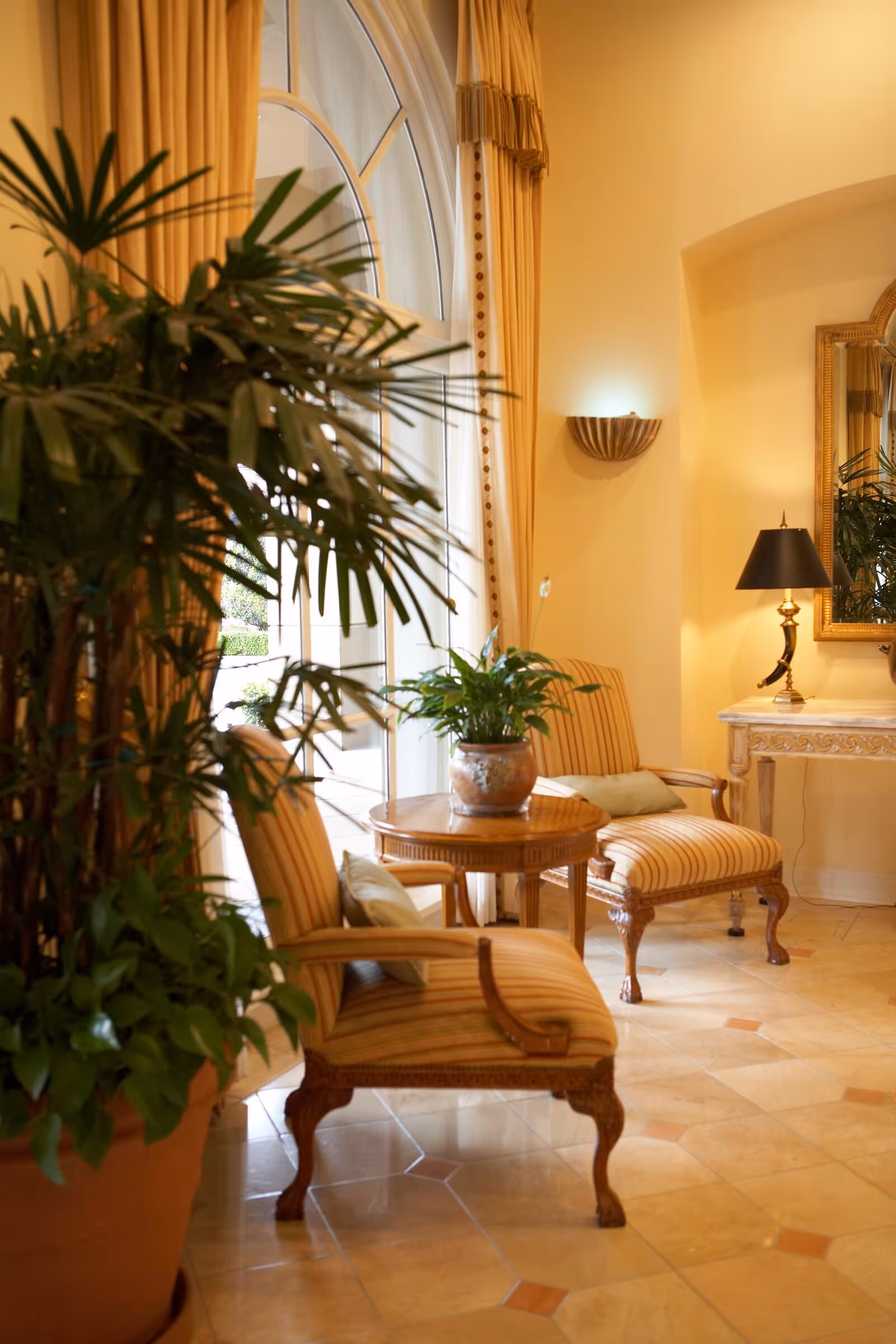 Sunlit elegant seating area with striped armchairs, a small round table, potted plants, and a lamp by an arched window.
