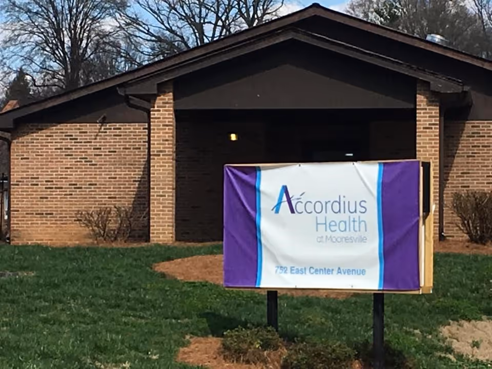 Exterior view of a brick building with a dark brown roof and a sign in front that reads 'Accordius Health at Mooresville, 752 East Center Avenue'. The area around the building has green grass and some small bushes.