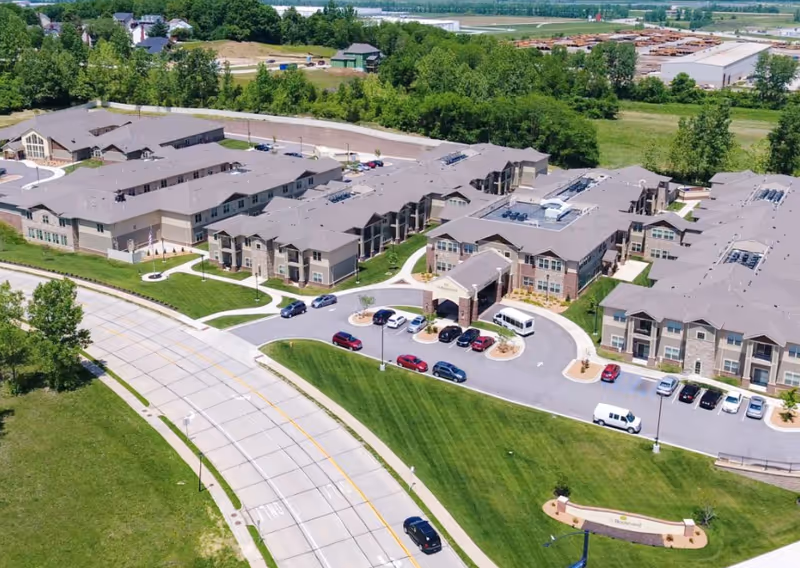 Aerial view of The Boulevard Senior Living Of Saint Charles facility showing multiple connected buildings with gray roofs, surrounded by green lawns and trees. There is a curved driveway with parked cars and a white van near the main entrance. The surrounding area includes roads, open fields, and some industrial buildings in the distance.