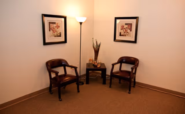 A small seating area with two dark wooden chairs with armrests placed on either side of a small black table. On the table is a decorative glass vase and some small ornamental items. A tall floor lamp with a white shade stands behind the table, illuminating the corner. Two framed floral artworks hang on the beige walls above each chair.