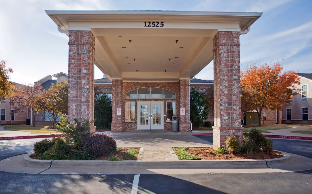 Front entrance of a senior living facility with a covered drop-off area supported by brick pillars, surrounded by landscaped bushes and trees with autumn foliage. The building number 12525 is displayed above the entrance doors.