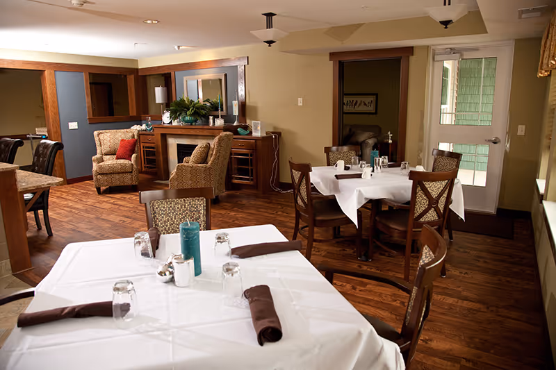 A cozy dining area with tables covered in white tablecloths, each set with glasses, napkins, and candles. The room features wooden flooring, comfortable upholstered chairs, and a sitting area with armchairs and a fireplace in the background. A door with glass panels leads to another space.