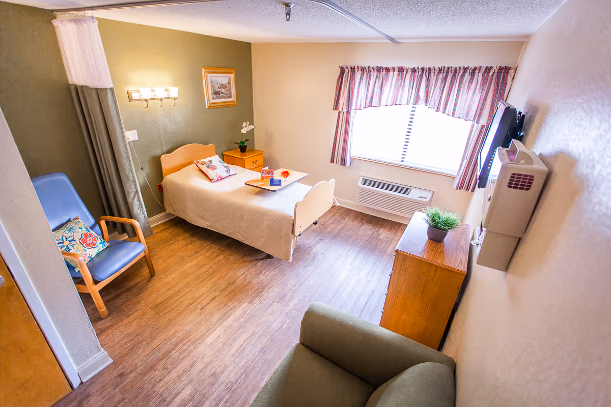 A small, cozy bedroom in a senior living facility with a single bed covered in a beige blanket and a floral pillow. There is a wooden nightstand with a small plant and a picture frame on the wall above it. A blue armchair with a colorful cushion is placed near the bed, and a green armchair is partially visible in the foreground. The room has wood flooring, a window with striped curtains, and a wall-mounted TV opposite the bed.