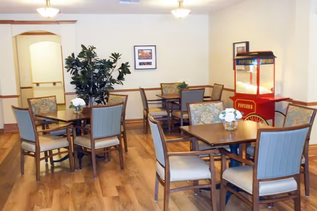 A dining area with several wooden tables and cushioned chairs arranged neatly on a wooden floor. Each table has a small floral centerpiece. There is a red popcorn machine in the corner and a large green plant near the back wall. The walls are light-colored with framed artwork and wooden trim.