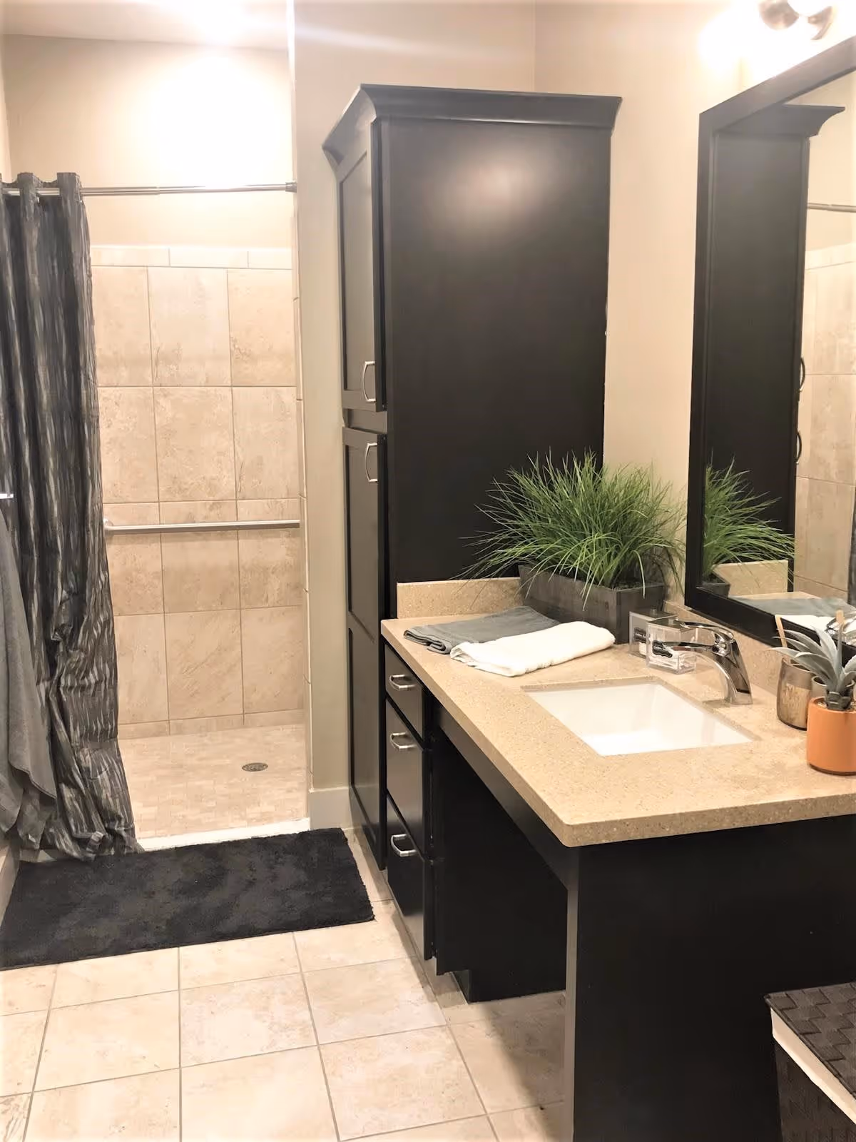 A bathroom with beige tiled floor and shower area with a curtain. There is a black cabinet with drawers and a beige countertop with a sink. On the countertop, there are folded towels and two potted plants. A large mirror is mounted on the wall above the sink.