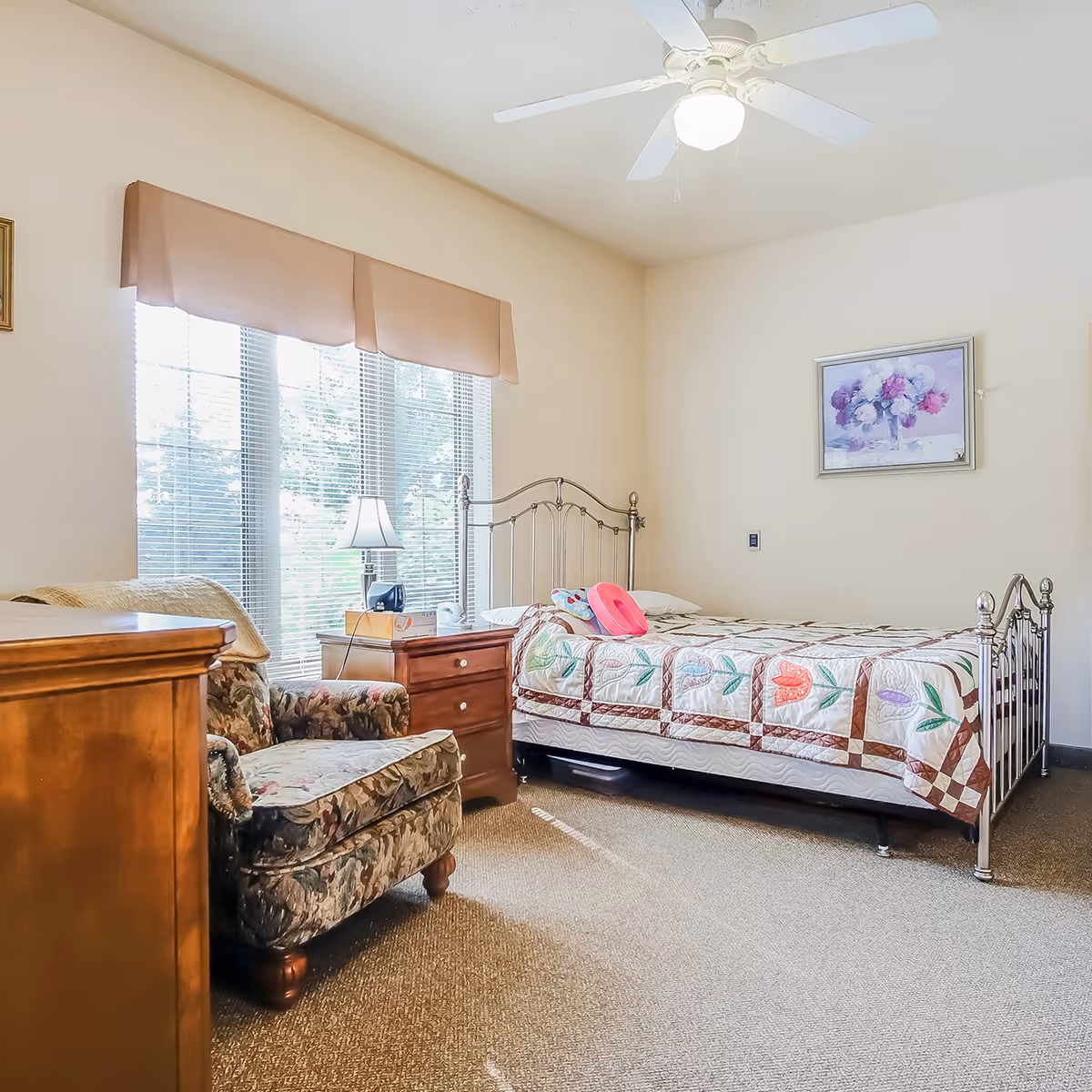 A bright and cozy bedroom in an assisted living facility featuring a single metal bed with a colorful quilt, a wooden nightstand with a lamp and phone, a floral upholstered armchair, a wooden dresser, and a ceiling fan. Large windows with blinds and a beige valance allow natural light to fill the room. A framed floral painting hangs on the wall.