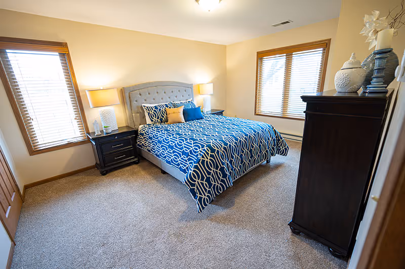 A bedroom with a large bed featuring a blue and white patterned comforter and multiple pillows. There are two windows with wooden blinds, a nightstand with a lamp on each side of the bed, and a dark wooden dresser with decorative vases on top. The room has beige walls and carpeted flooring.