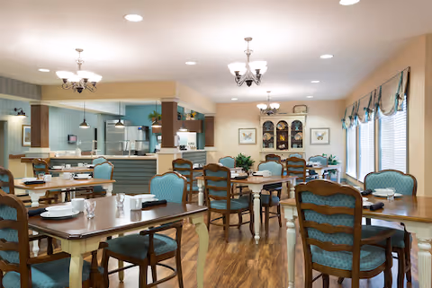 Dining room with multiple wooden tables and teal-upholstered chairs, hardwood floor, and a serving counter in the background.