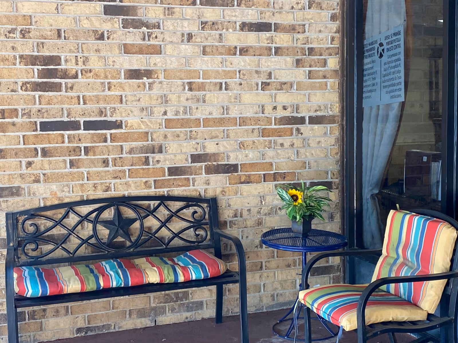 Metal bench and a striped-cushion chair flanking a small blue table with a potted sunflower, set against a brick wall and glass door.