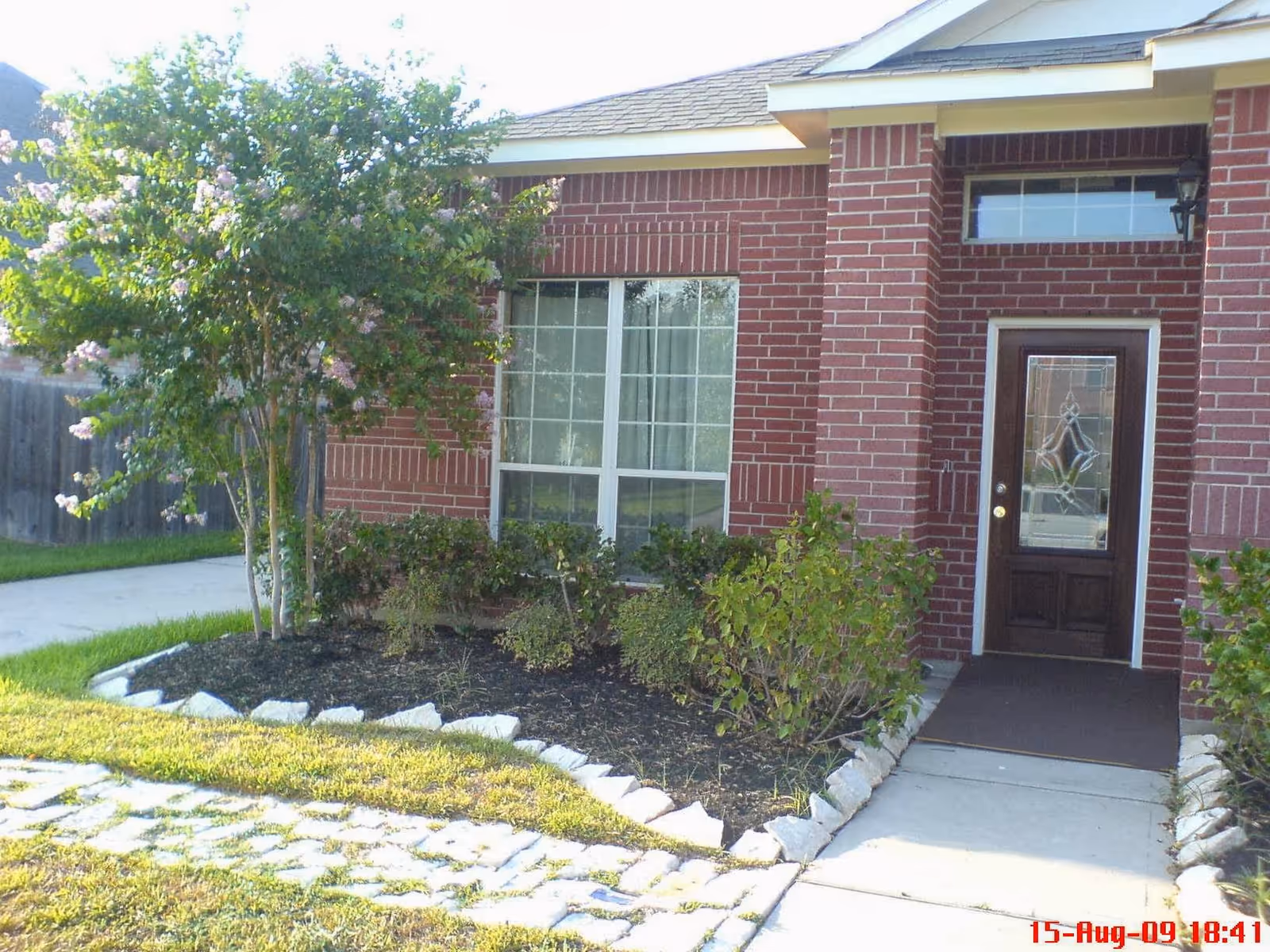 Front entrance of a brick house with a glass-panel door, large window, small landscaped garden and a paved walkway.
