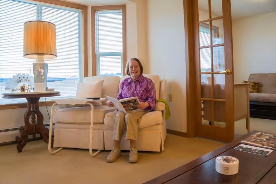 An elderly woman sitting on a beige sofa in a well-lit living room, holding an open book and smiling. The room features large windows with blinds, a wooden side table with a lamp and decorative items, a glass-paned wooden door, and a coffee table with magazines and a small container.