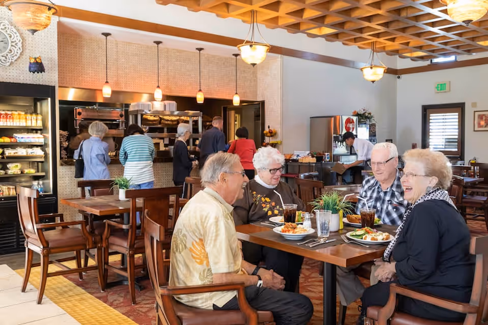 A group of four elderly people sitting around a wooden dining table in a well-lit dining area, enjoying a meal and conversation. In the background, several other people are standing near a food service counter with hanging pendant lights, and a staff member is working near a beverage machine. The room features wooden ceiling beams, warm lighting, and a cozy atmosphere.