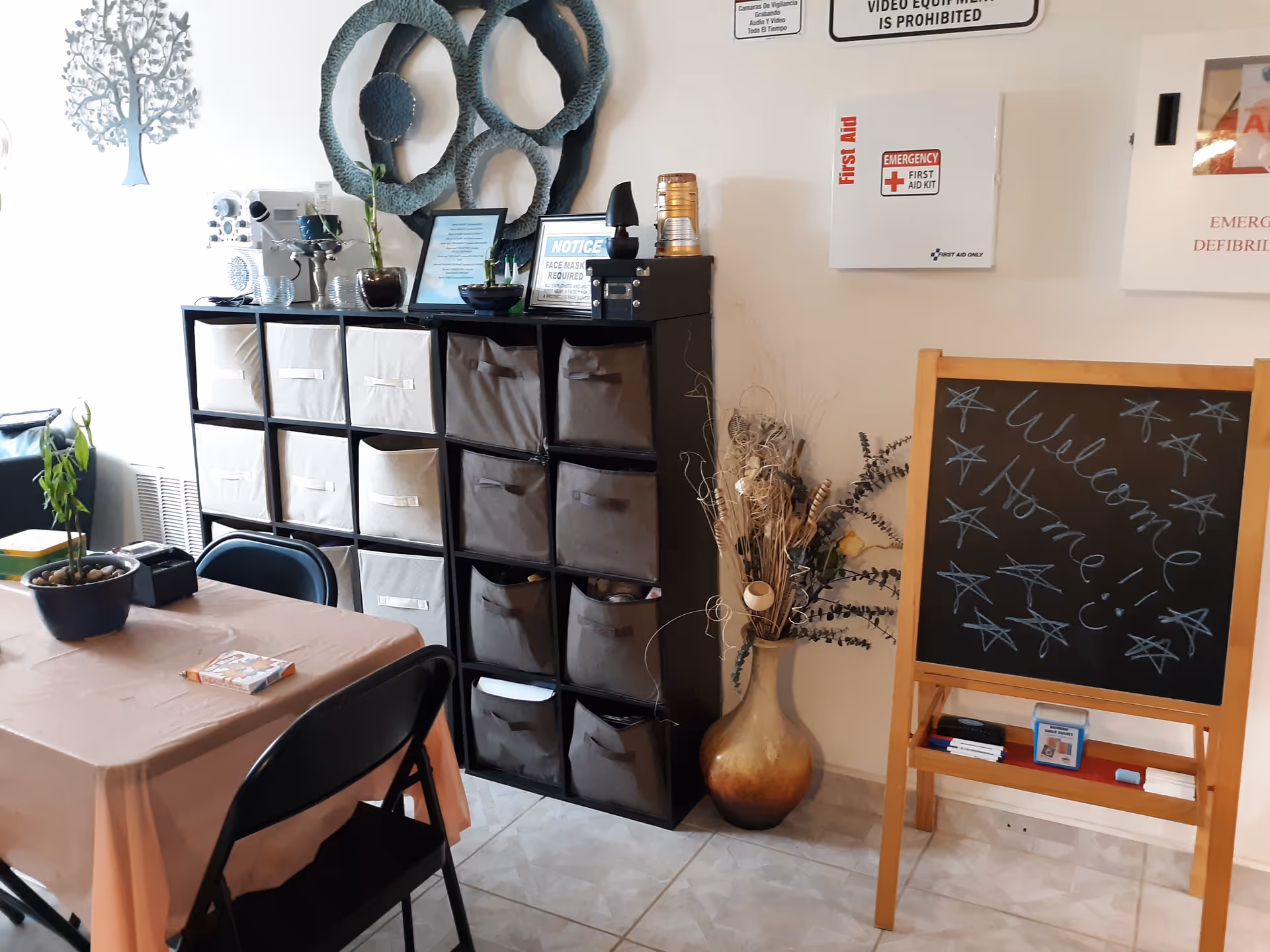 A communal room with a table and folding chairs, cube storage units, decorative vase, and a chalkboard that says "Welcome Home".