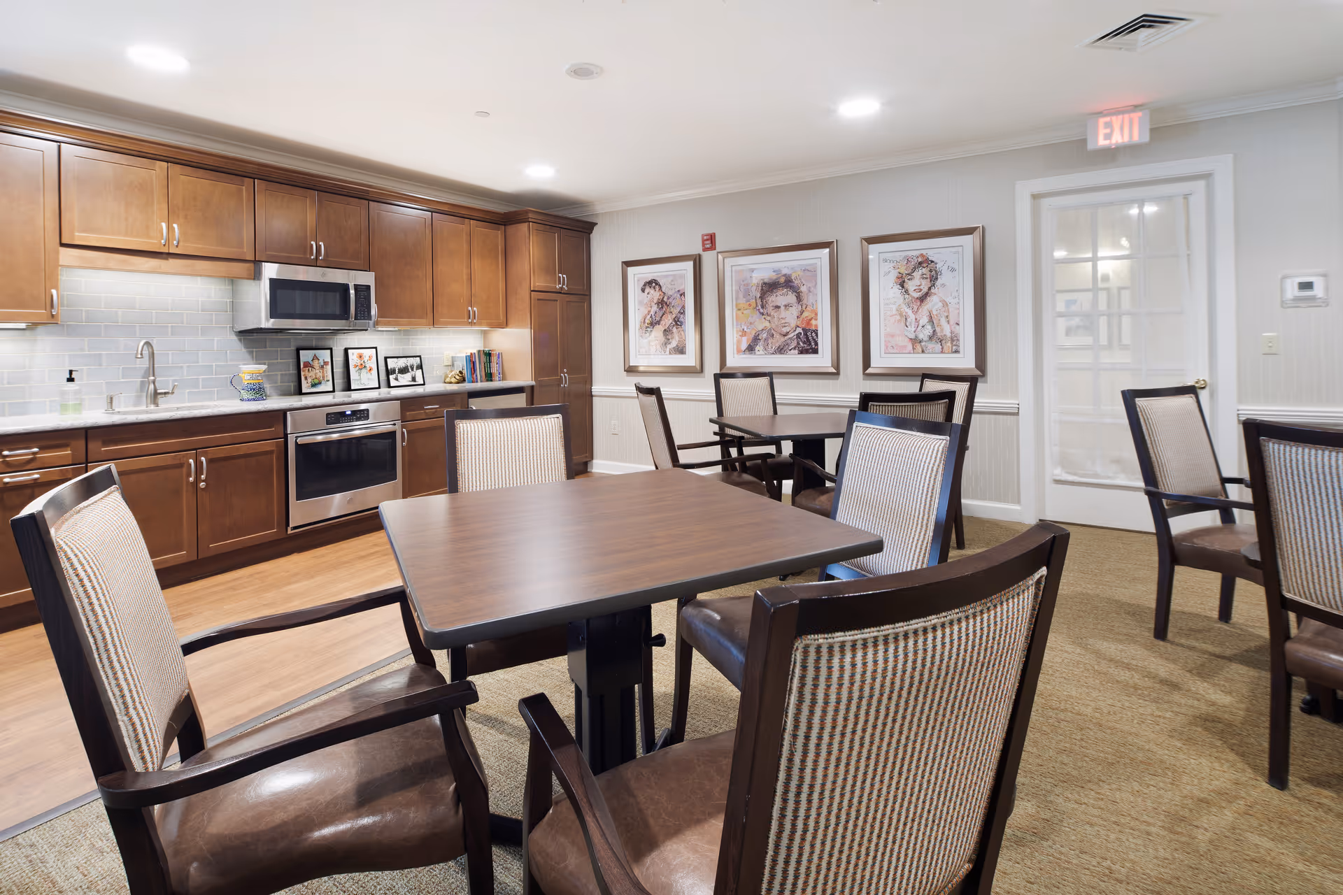A well-lit communal kitchen and dining area with wooden cabinets, stainless steel microwave and oven, and several tables surrounded by cushioned chairs. The walls are decorated with framed artwork, and there is an exit door with a glass panel and an illuminated exit sign above it.