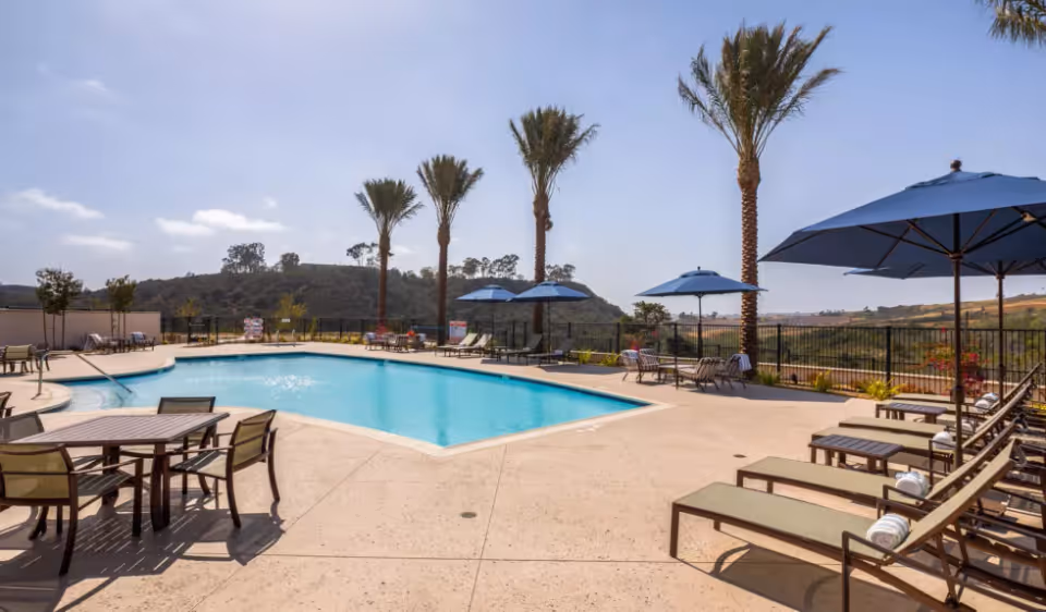 Outdoor swimming pool area with lounge chairs, tables, blue umbrellas and palm trees overlooking hills.
