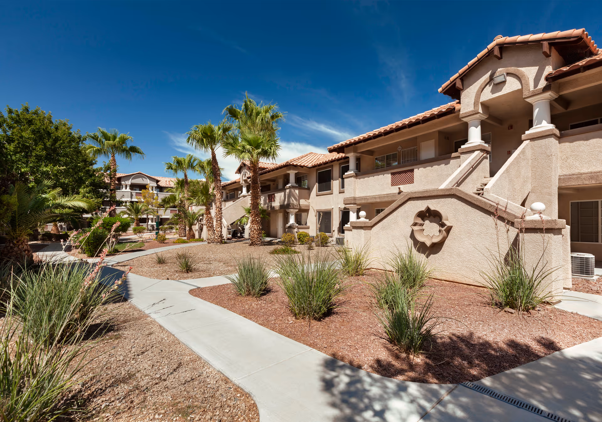 Exterior view of a residential building at Destinations Pebble with beige stucco walls, red tile roof, palm trees, and landscaped desert plants under a clear blue sky.