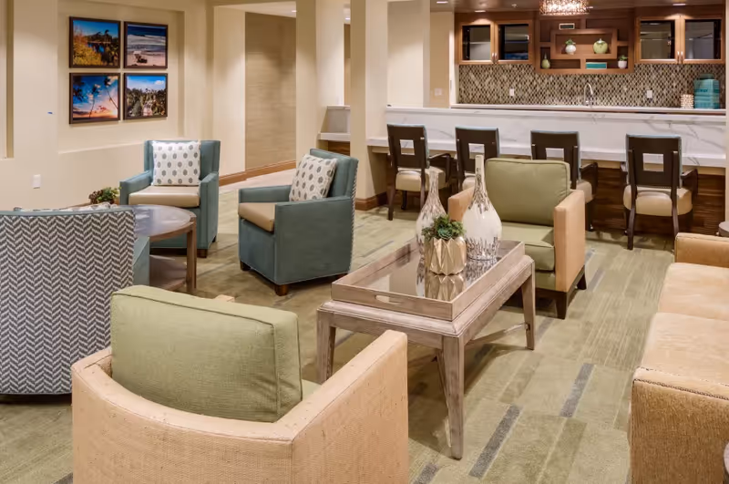 Communal lounge with upholstered chairs, a central coffee table with decorative vases, and a bar counter with stools.