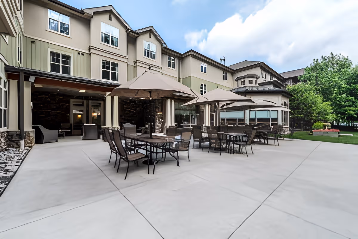 Outdoor patio area at Spring Mill facility with multiple tables and chairs under large beige umbrellas, adjacent to a multi-story building with windows and surrounded by greenery.