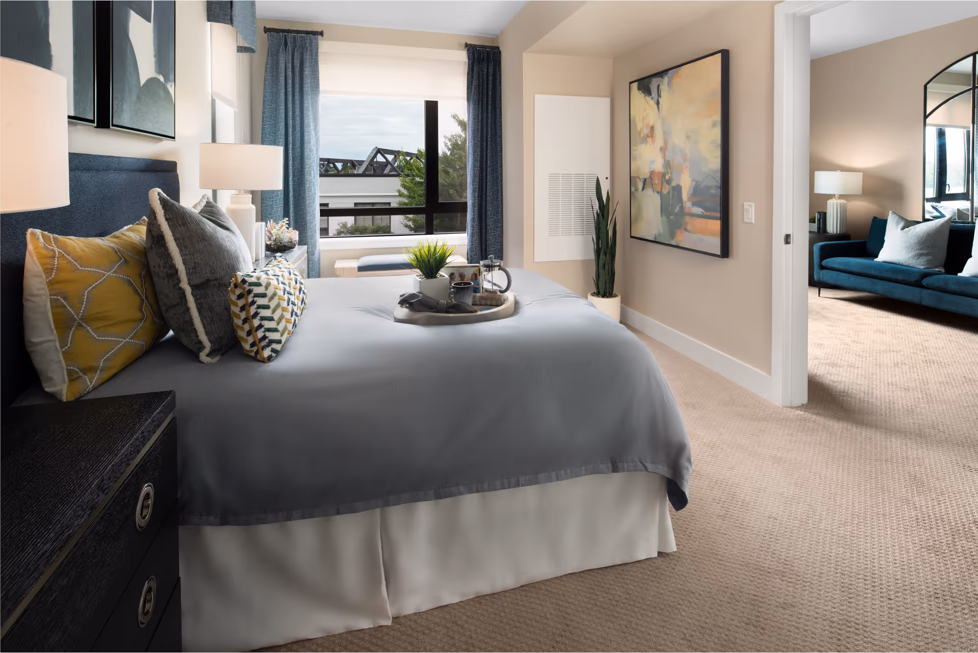 A cozy bedroom with a neatly made bed featuring gray and yellow pillows and a gray bedspread. A tray with a plant, a cup, and a photo frame is placed on the bed. There is a window with blue curtains letting in natural light, a large abstract painting on the wall, and a potted plant in the corner. Through an open doorway, a living room with a teal sofa, pillows, a lamp, and a large mirror is visible.