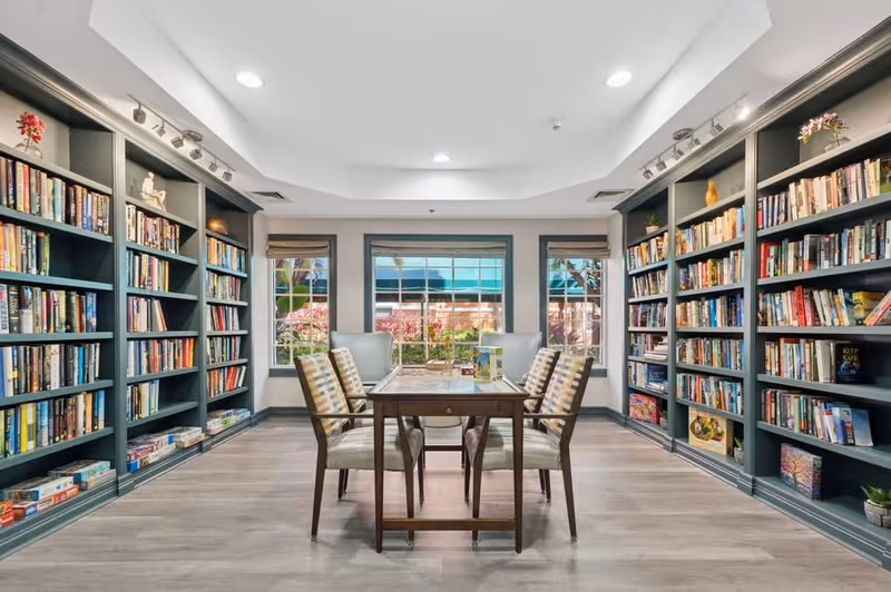 A bright and spacious library room with large windows showing outdoor greenery. The room features built-in bookshelves filled with books and board games on both sides. In the center, there is a wooden table with six cushioned chairs arranged around it, suitable for reading or playing games.