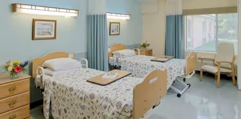 Two hospital-style beds in a shared care room with blue curtains and furnishings.