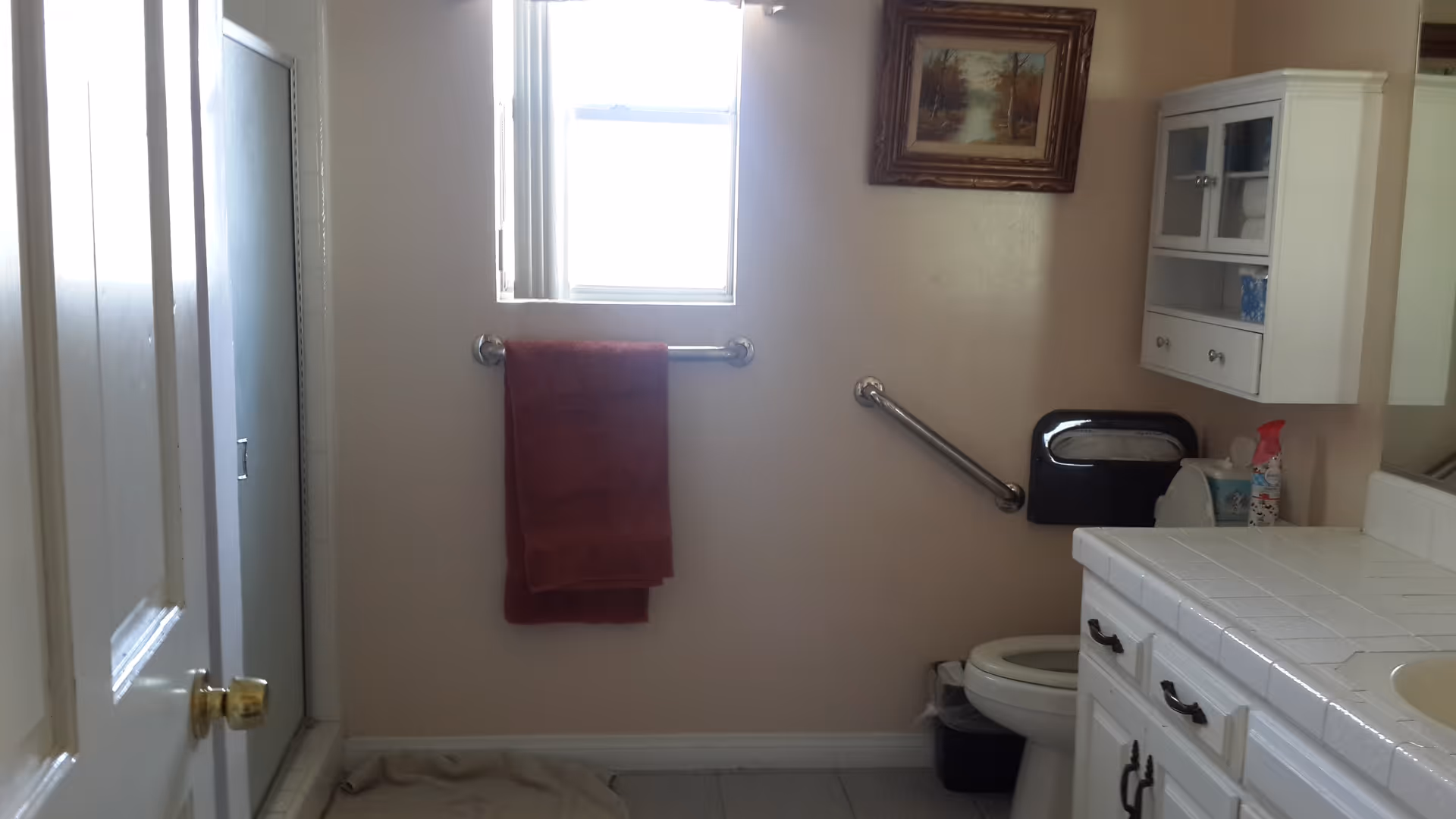 A bathroom with a white tiled countertop and sink on the right, a toilet with a grab bar beside it, a small wall-mounted cabinet above the toilet, a red towel hanging on a towel bar under a window, and a framed picture on the wall.