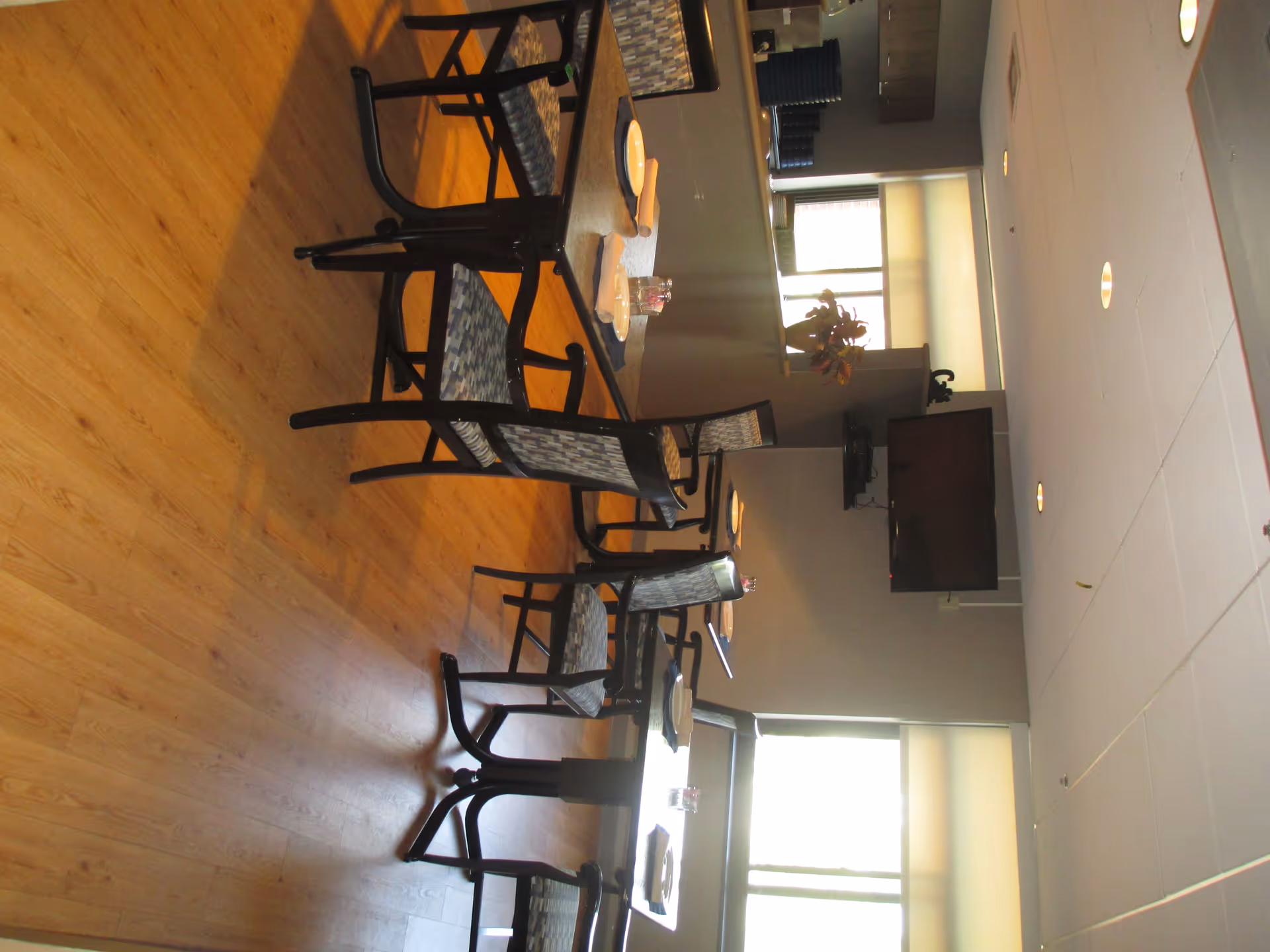 Interior view of a dining area with wooden flooring, several tables set with plates and glasses, and chairs with patterned cushions. A large window with blinds allows natural light into the room, and a flat-screen TV is mounted on the wall.