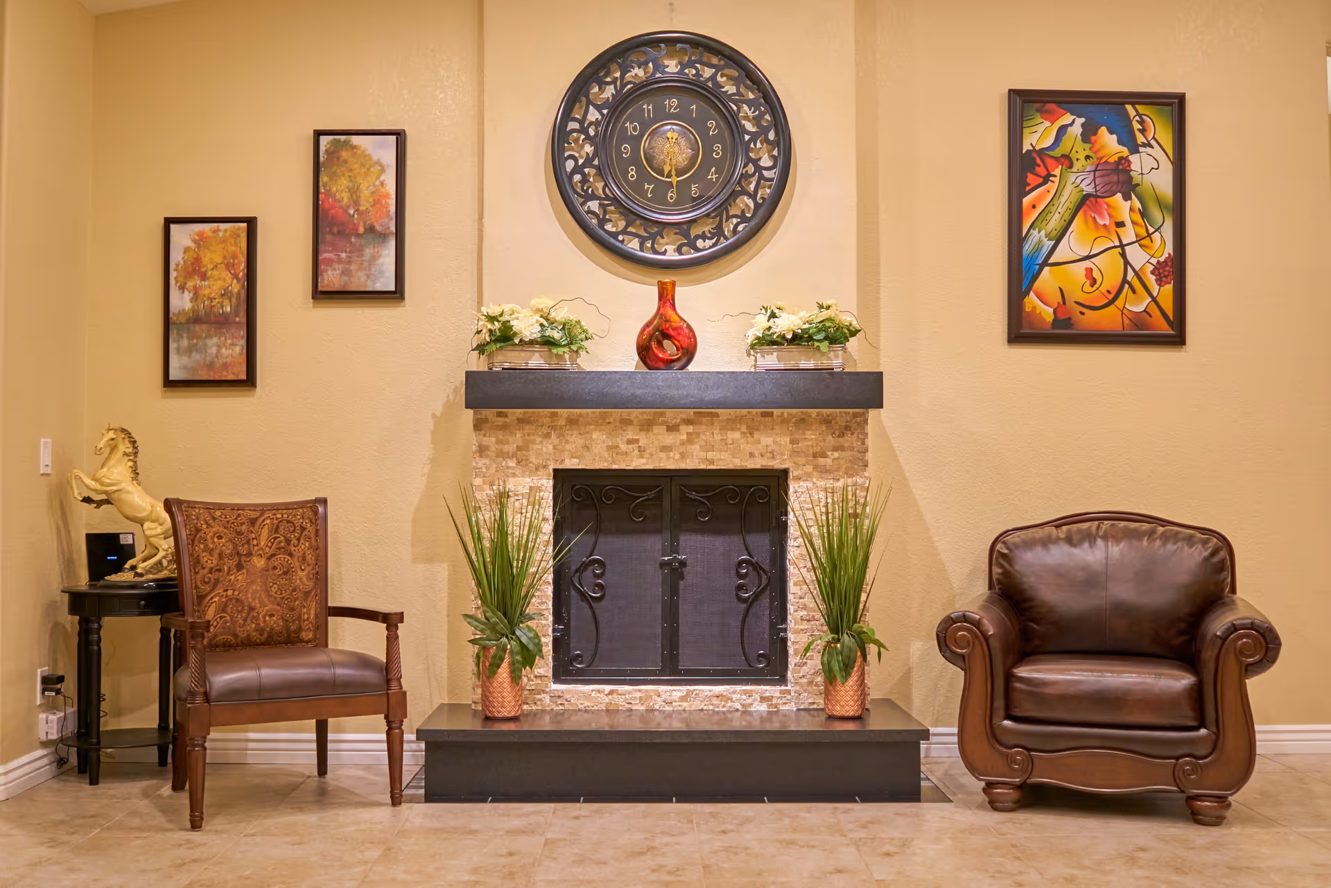 Interior seating area with a central fireplace, decorative clock and artwork, flanked by two chairs and potted plants.