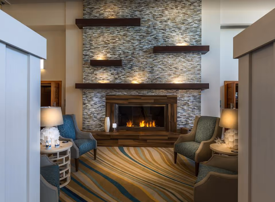 A cozy living room area with a modern fireplace set in a tiled wall with wooden shelves above it. The room features four upholstered armchairs arranged around the fireplace, two on each side, with side tables and lamps providing warm lighting. The floor has a patterned carpet with flowing lines in shades of beige, blue, and brown.