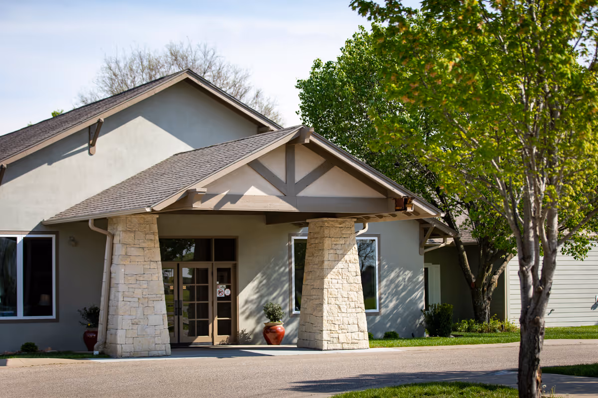 Exterior view of a building entrance with a peaked roof supported by stone columns, surrounded by trees and greenery under a clear sky.