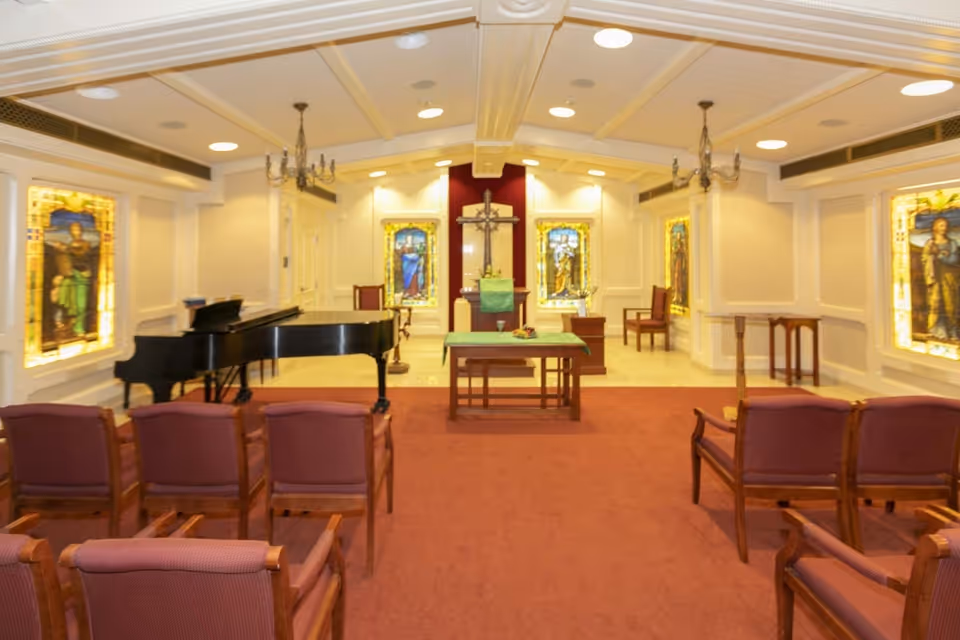 Interior of a chapel with rows of wooden chairs with red cushions facing an altar. The altar has a green cloth and a cross mounted on the wall behind it. There are stained glass windows on the walls and a black grand piano to the left side of the room. The ceiling has recessed lighting and chandeliers.