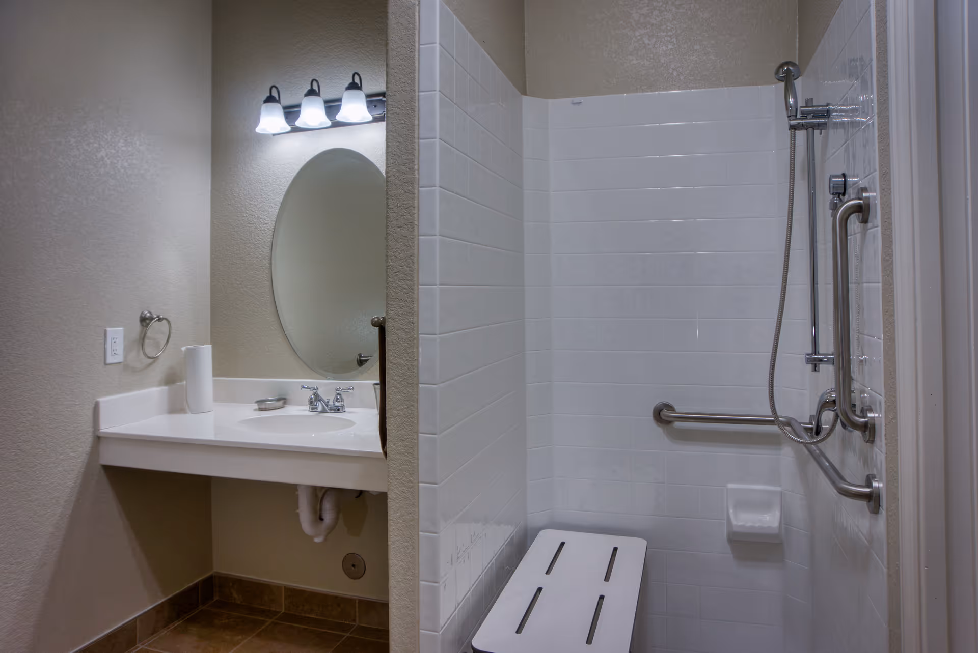 A bathroom with a white sink and countertop on the left, an oval mirror above the sink, and a three-light fixture. On the right, there is a tiled shower area with a handheld showerhead, grab bars, and a white shower bench.