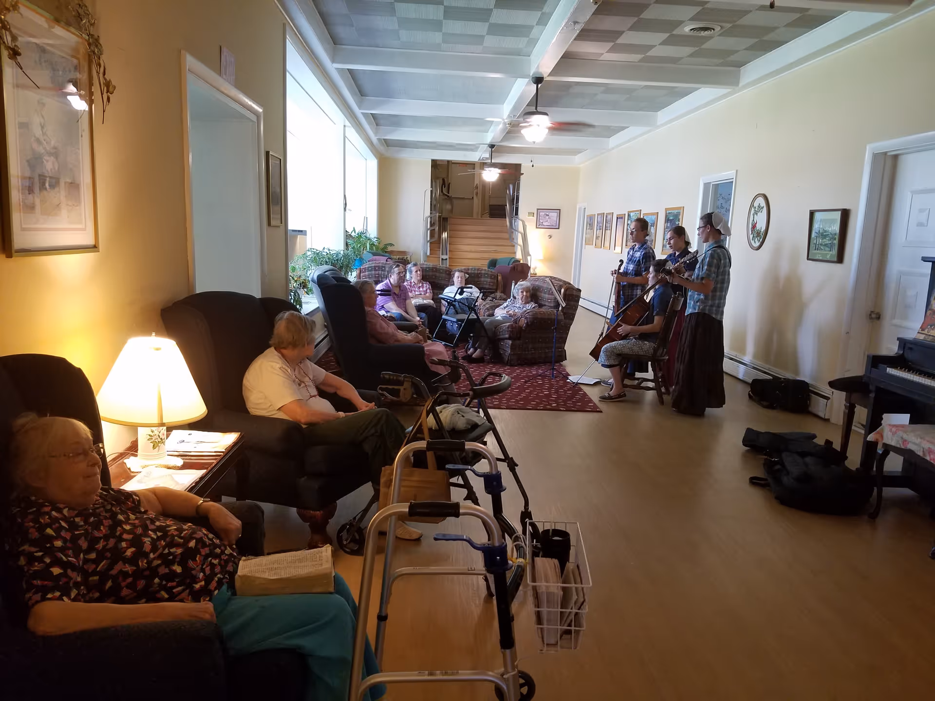 A group of elderly people sitting in armchairs and sofas in a long, well-lit living room with wooden floors. Some are watching a small group of musicians playing string instruments near the far wall. Walkers are visible in the foreground, and framed pictures hang on the walls.