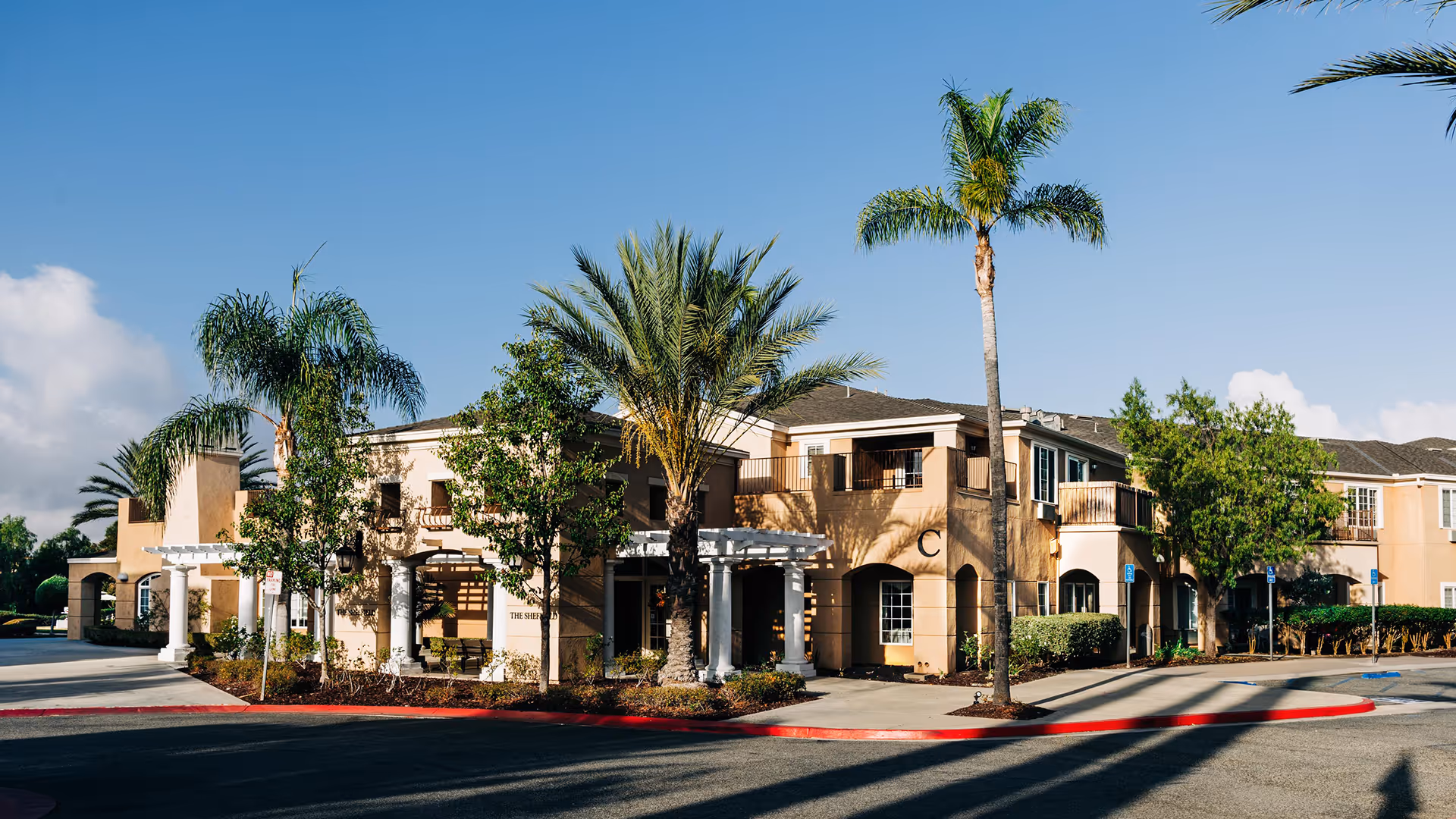 Exterior view of a two-story senior living facility building with beige walls, white columns, and several palm trees and other greenery around the entrance. The building has balconies and multiple windows under a clear blue sky.