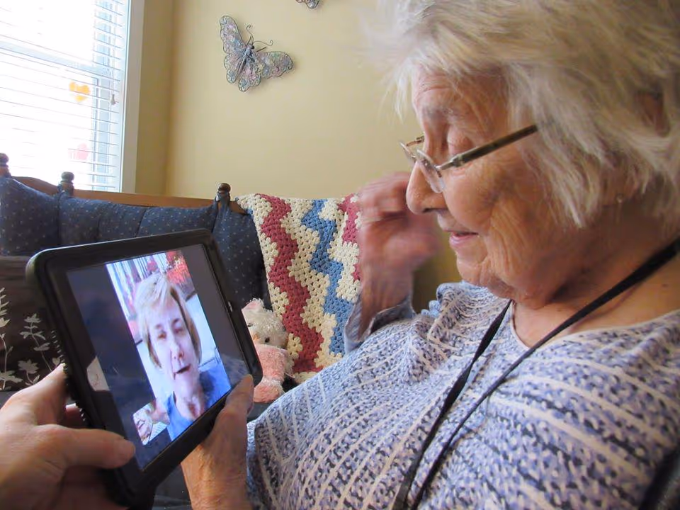 An elderly woman with glasses is sitting on a couch with a crocheted blanket and pillows, smiling while video chatting on a tablet held by another person. A butterfly decoration is visible on the wall behind her.