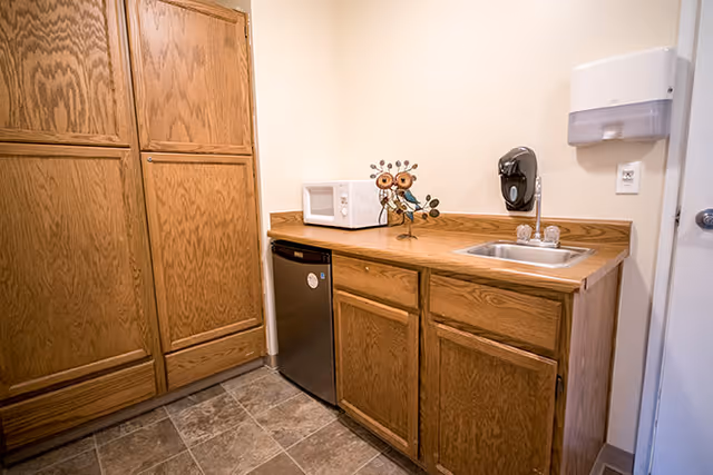 Small kitchenette area with wooden cabinets, a mini refrigerator, a microwave, a decorative owl figurine, a stainless steel sink, and a wall-mounted soap dispenser.