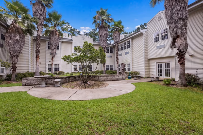 Courtyard with palm trees, a circular concrete walkway and benches in front of a light-colored multi-story assisted living building.