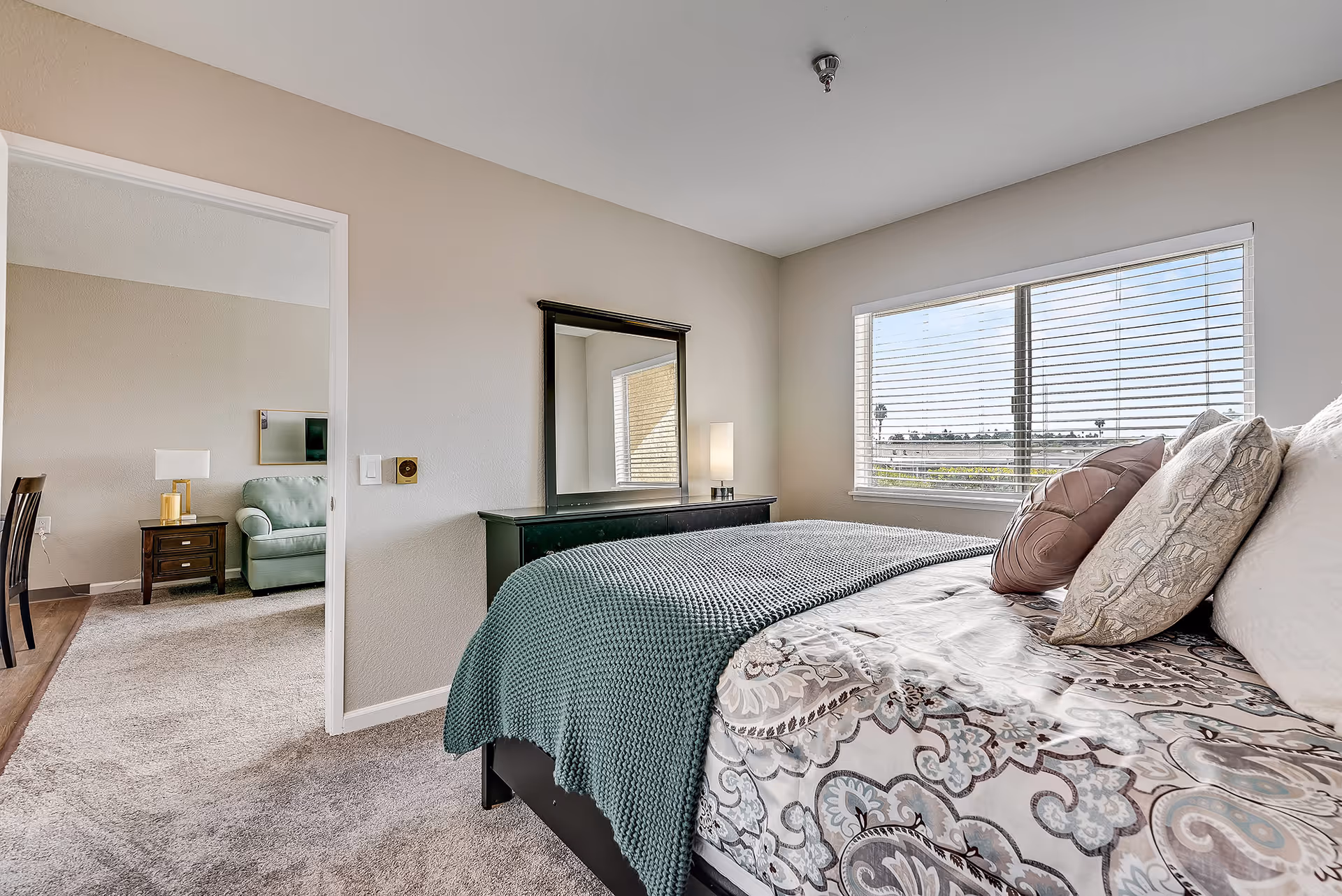 A bright bedroom with a large window covered by blinds, a bed with patterned bedding and multiple pillows, a dark dresser with a mirror and a lamp, and an open doorway leading to a living area with a green armchair and a side table with a lamp.