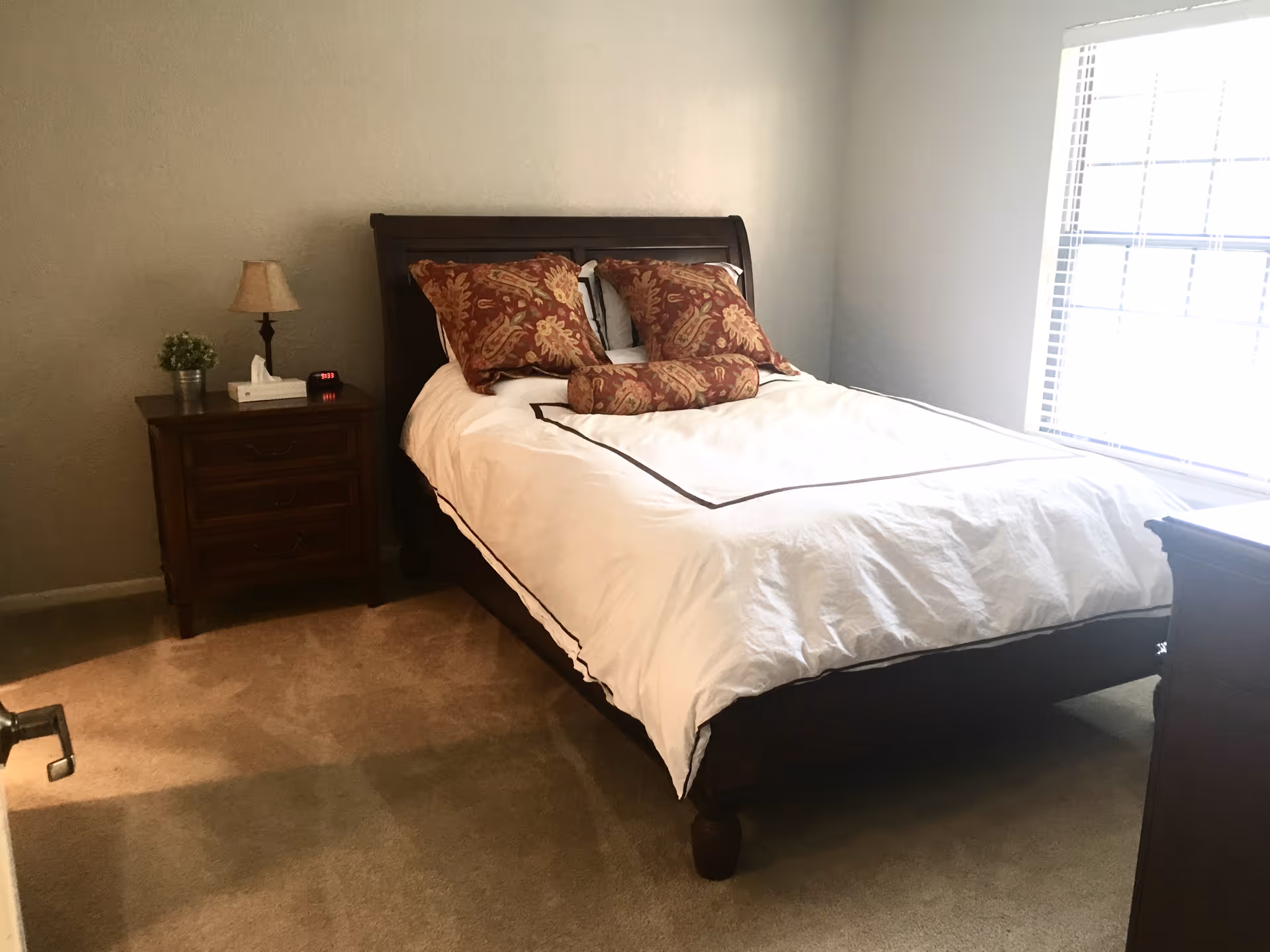 A neatly made bed with white bedding and patterned pillows in a bright bedroom with a wooden nightstand and window.