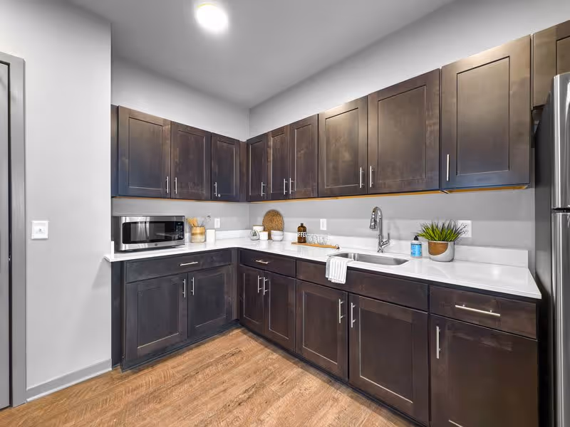 Modern kitchen with dark wood cabinets, white countertops, a stainless steel refrigerator, a microwave, a sink with a faucet, and various kitchen items including a plant, a coffee container, and utensils.